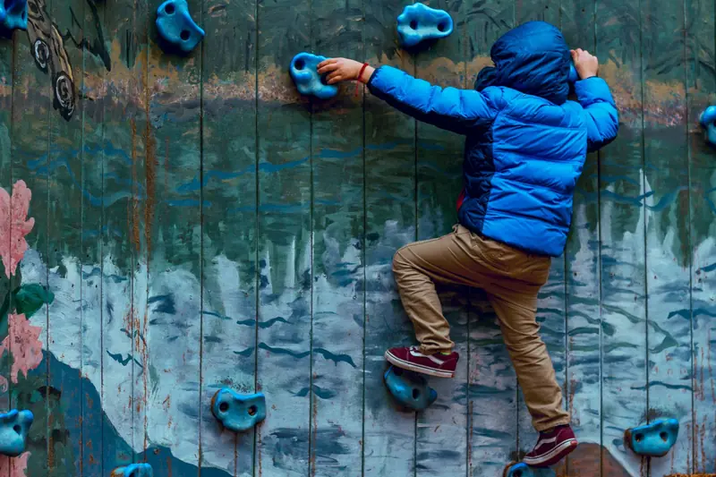 Young boy in blue jacket climbing a painted rock wall outdoors, showcasing adventure and play