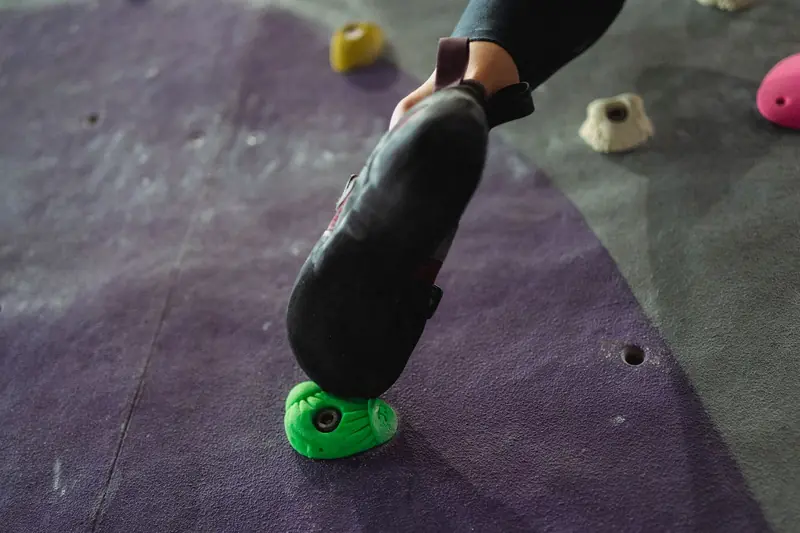 From below crop climber in leggings and sneakers putting foot on climbing wall holder in modern fitness club
