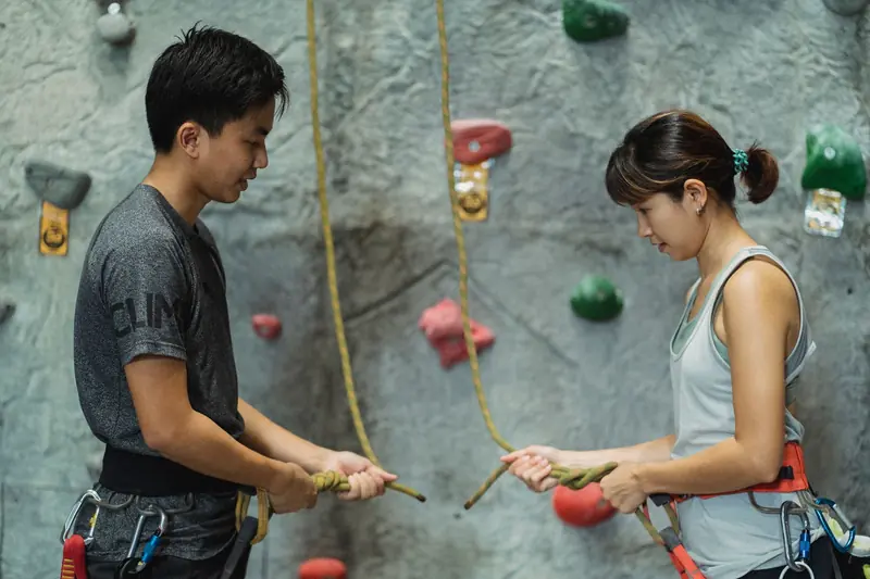 Side view of young ethnic male instructor showing to female climber how to tie safety rope near climbing wall