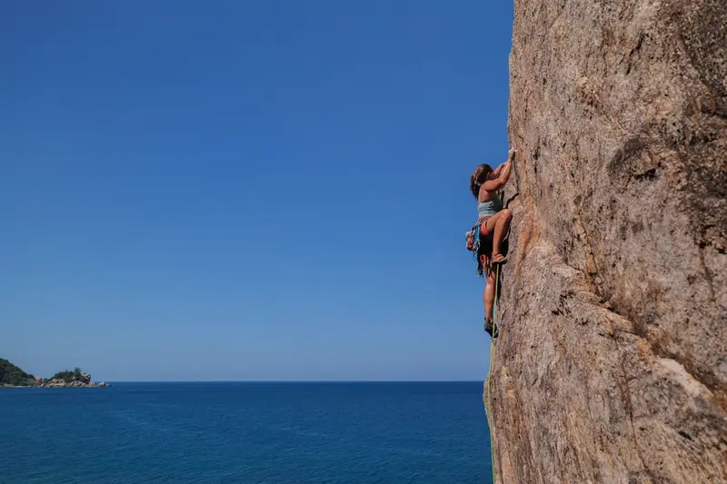 A woman skillfully rock climbing a steep cliff with a clear blue sky and sea view