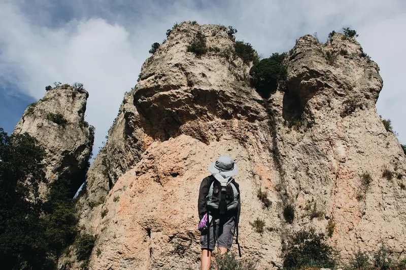 Man with backpack stands before a dramatic rock formation under a bright blue sky, suggesting adventure and exploration
